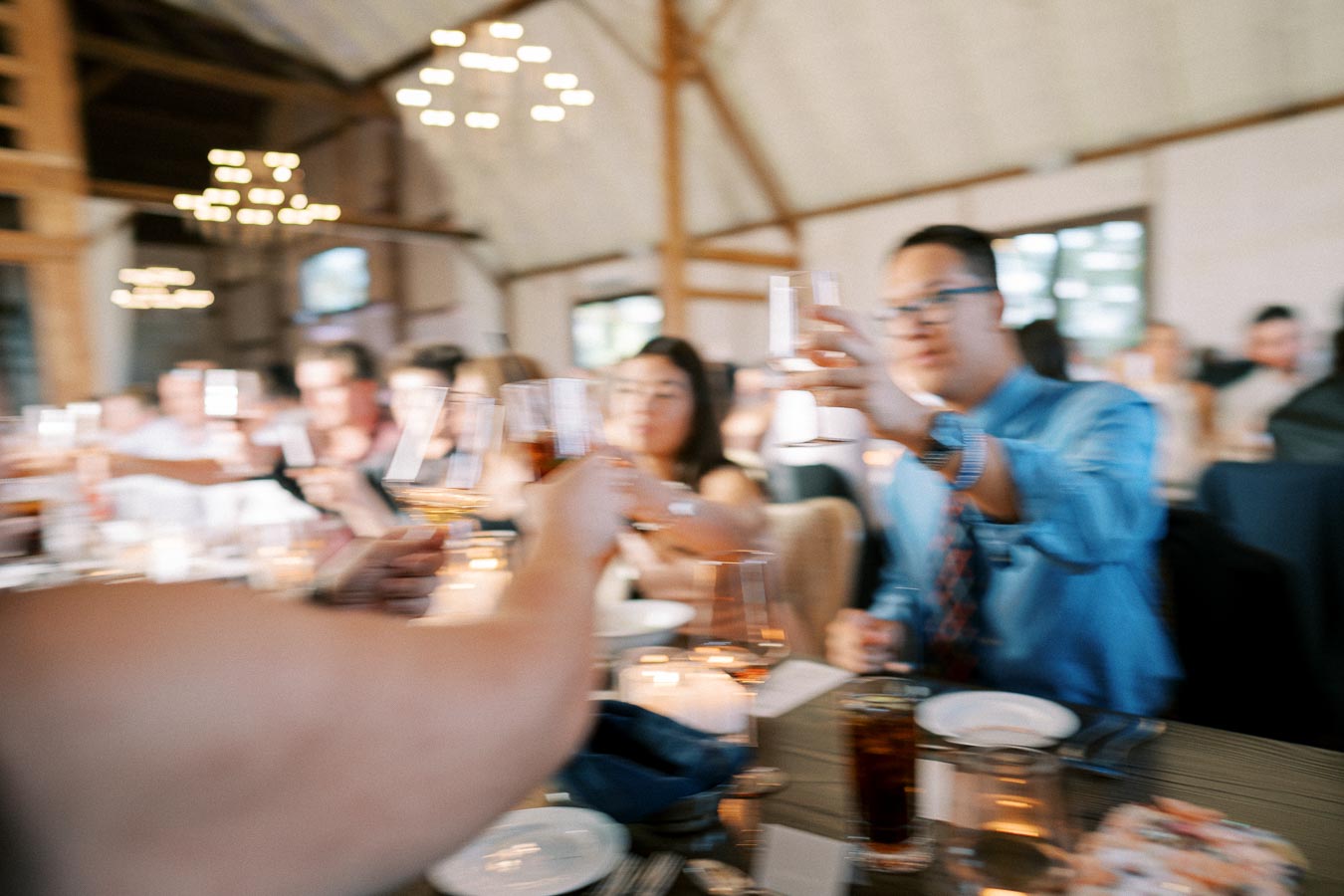 A group of people at a celebratory event raising glasses in a toast in a warmly lit venue with chandeliers and wooden beams.