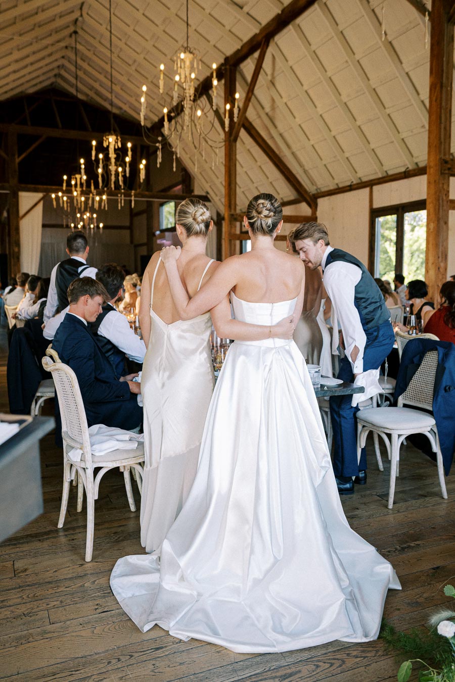 Two brides in elegant white gowns embrace during their wedding reception in a rustic barn setting. Guests are seated at beautifully decorated tables with ambient lighting from chandeliers.