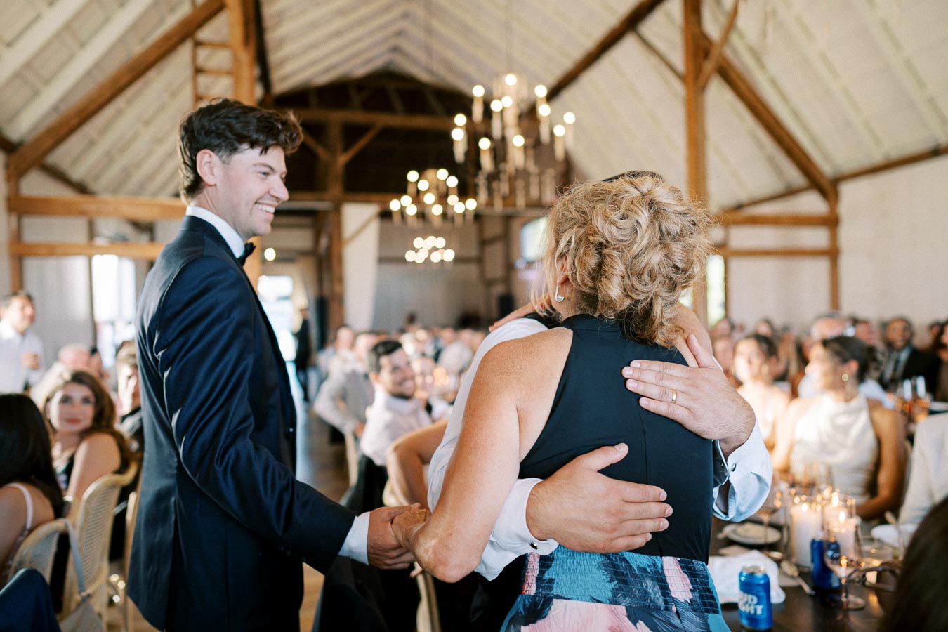A joyful wedding reception moment with a man in a suit greeting guests, inside a beautifully decorated barn setting with chandeliers and wooden beams.