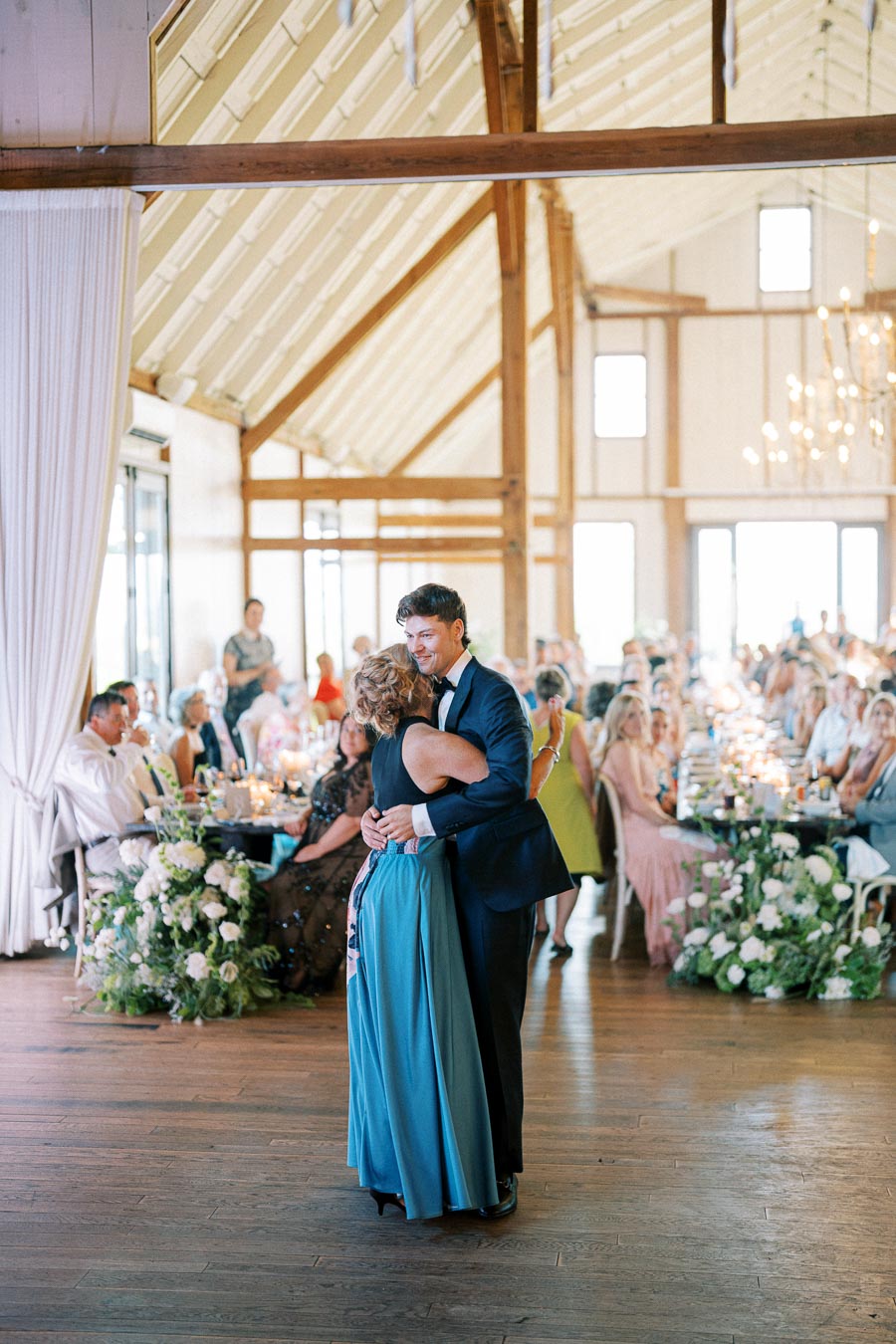 A groom in a suit dances with a woman in a blue dress during a wedding reception in a beautifully decorated barn, surrounded by guests seated at elegantly set tables.