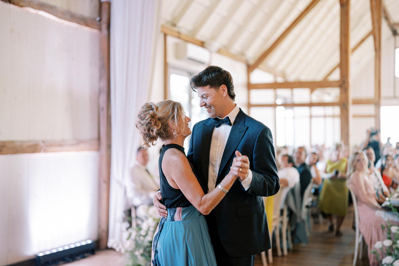 A couple sharing a joyful dance at a wedding reception inside a rustic venue, with guests seated in the background.