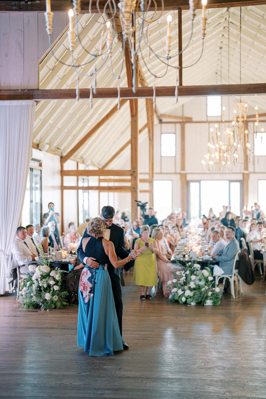 Couple dancing at a wedding reception inside a beautifully decorated barn venue, surrounded by seated guests and elegant chandeliers.