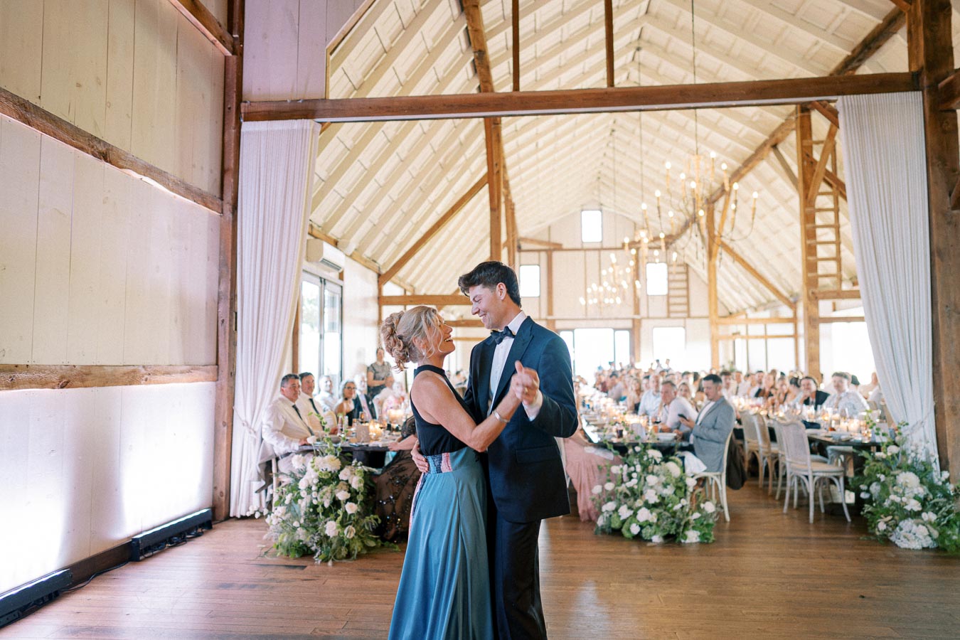 A couple enjoys a dance in a beautifully decorated rustic barn wedding venue, surrounded by seated guests and elegant floral arrangements.