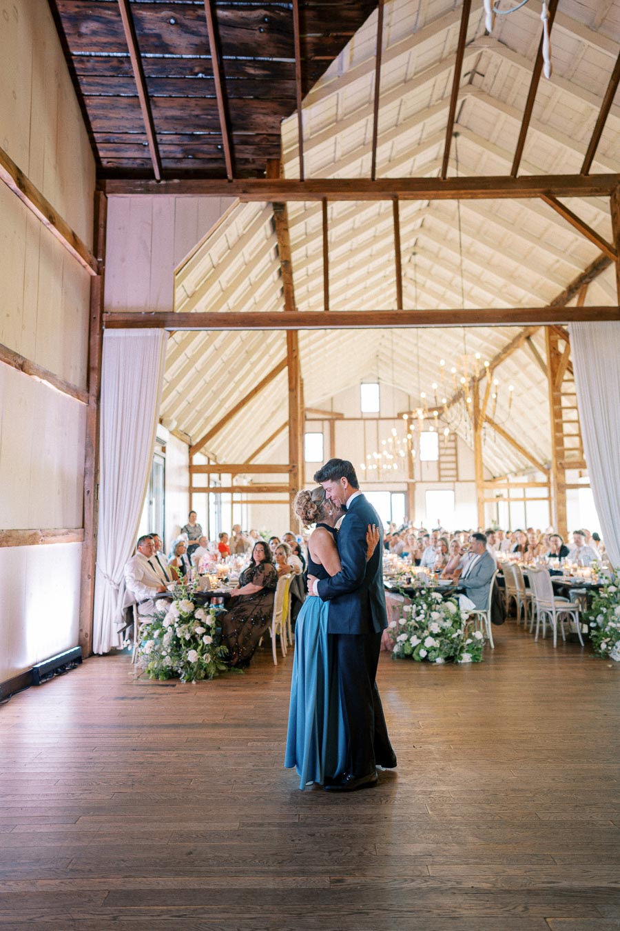 A couple sharing a dance in a beautifully decorated rustic barn during a wedding reception, surrounded by seated guests and elegant floral arrangements.