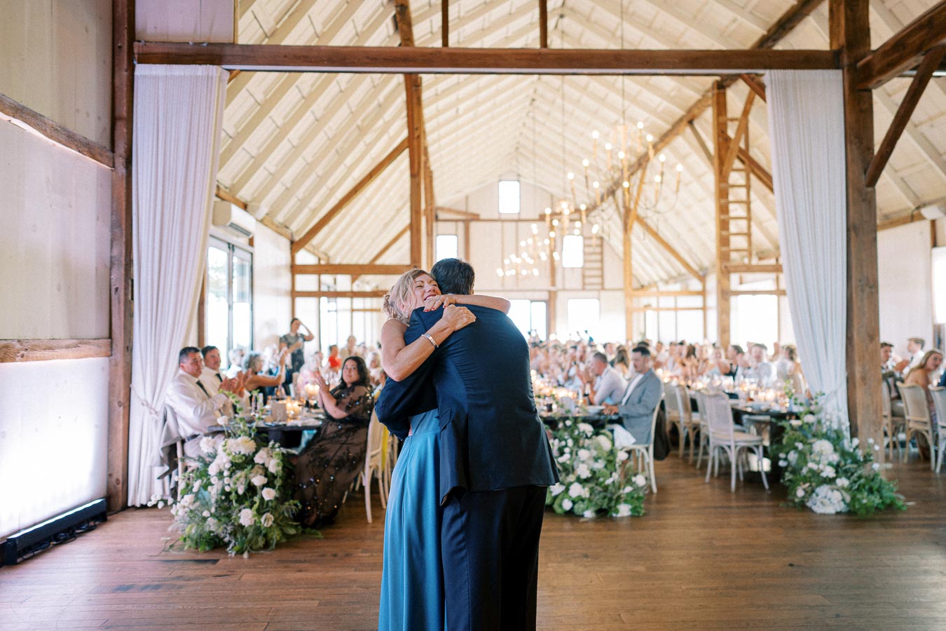 A couple embracing during a dance at a wedding reception in a rustic barn venue, with guests seated at elegantly decorated tables and floral arrangements lining the aisle.
