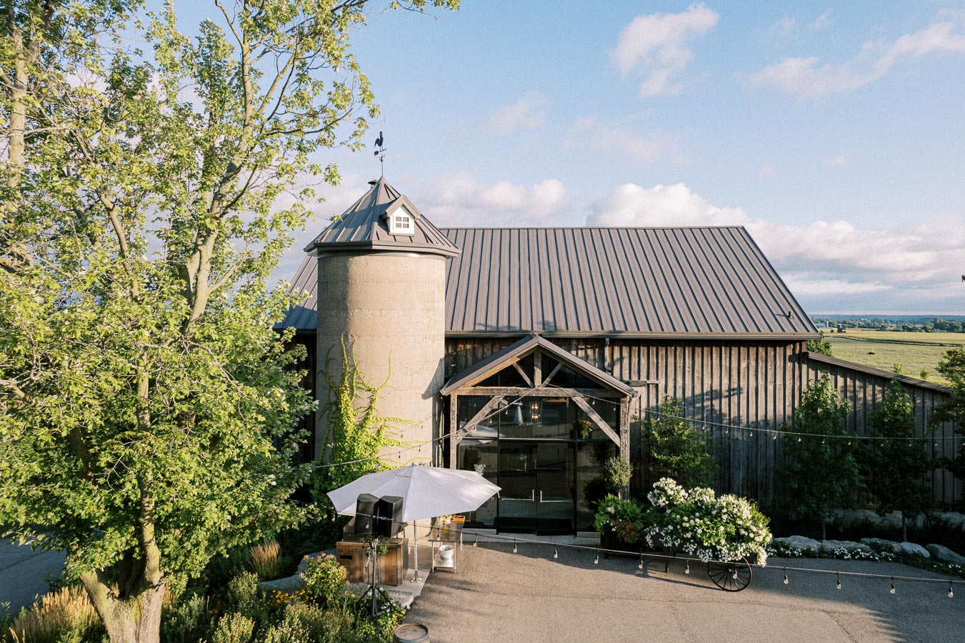 Scenic view of a rustic barn with a silo surrounded by lush greenery and fields, featuring a wooden entrance and outdoor patio area under a clear blue sky.