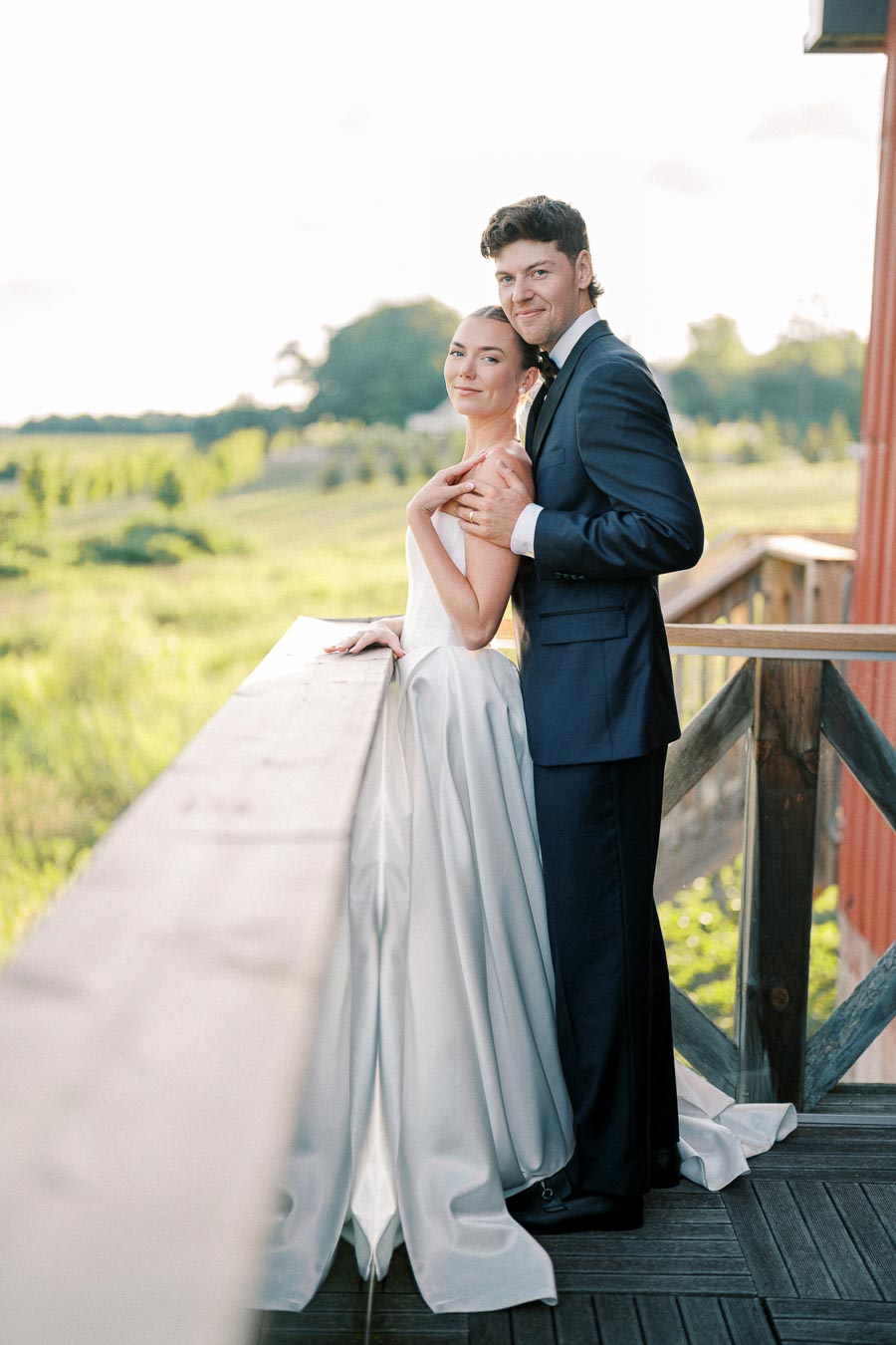 A bride in a white wedding dress and a groom in a dark suit smiling and embracing on a wooden balcony overlooking a lush green landscape.