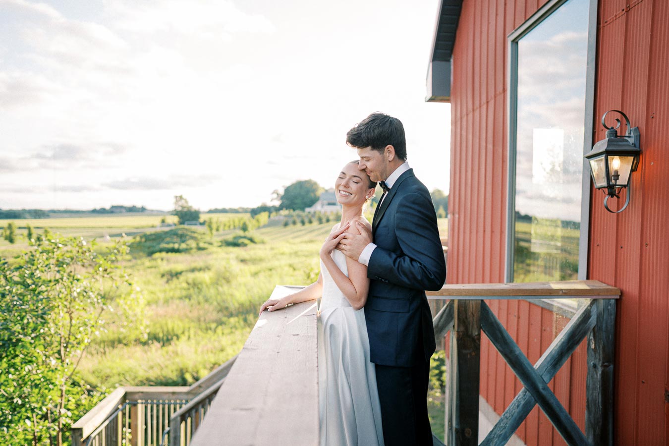 A bride in a white dress and groom in a dark suit embrace on a sunny balcony overlooking a lush green landscape and red building.