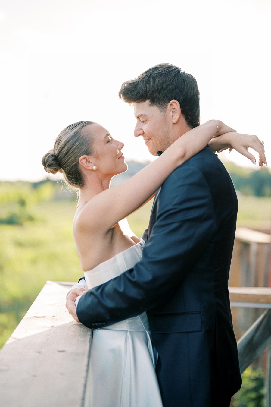 A couple embracing joyfully in a scenic outdoor setting, with the woman in a stylish white dress and the man in a sleek dark suit, capturing a romantic moment against a backdrop of lush greenery.