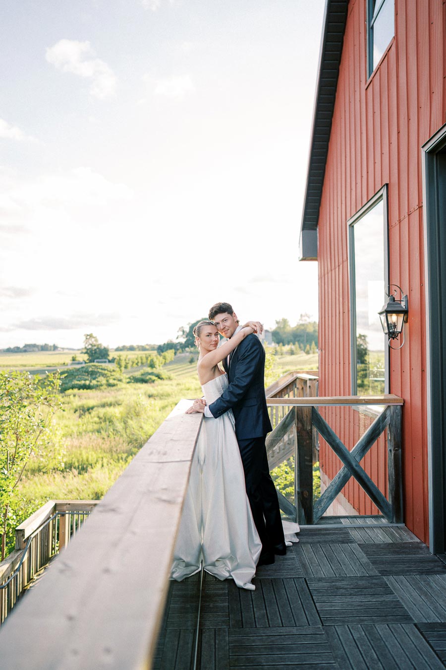 Bride and groom embracing on a rustic wooden balcony with scenic countryside backdrop, next to a red barn under a clear sky.