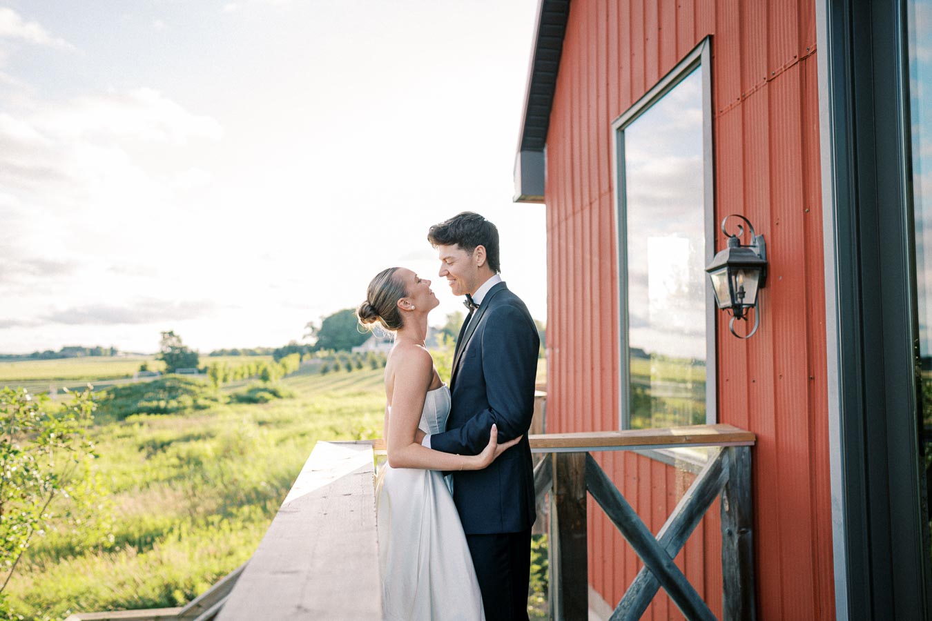 Bride and groom embracing on a sunny balcony with a scenic countryside backdrop, dressed in wedding attire next to a red barn-style building.