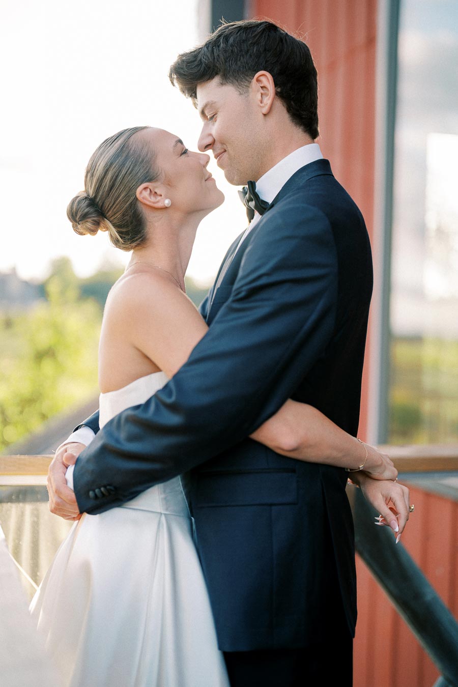Elegant couple embracing at sunset in wedding attire, bride in elegant white gown and groom in dark suit, romantic outdoor setting.