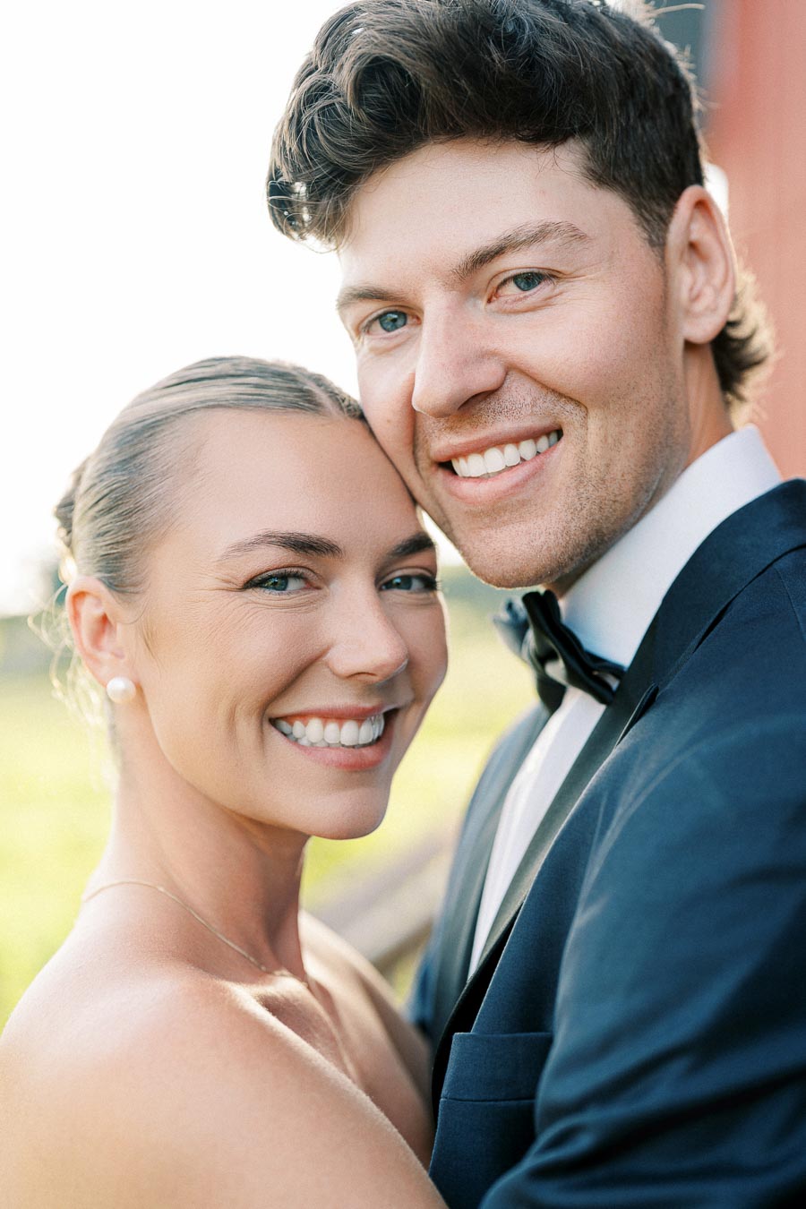 Smiling couple embracing outdoors, wearing elegant attire, against a sunny backdrop.