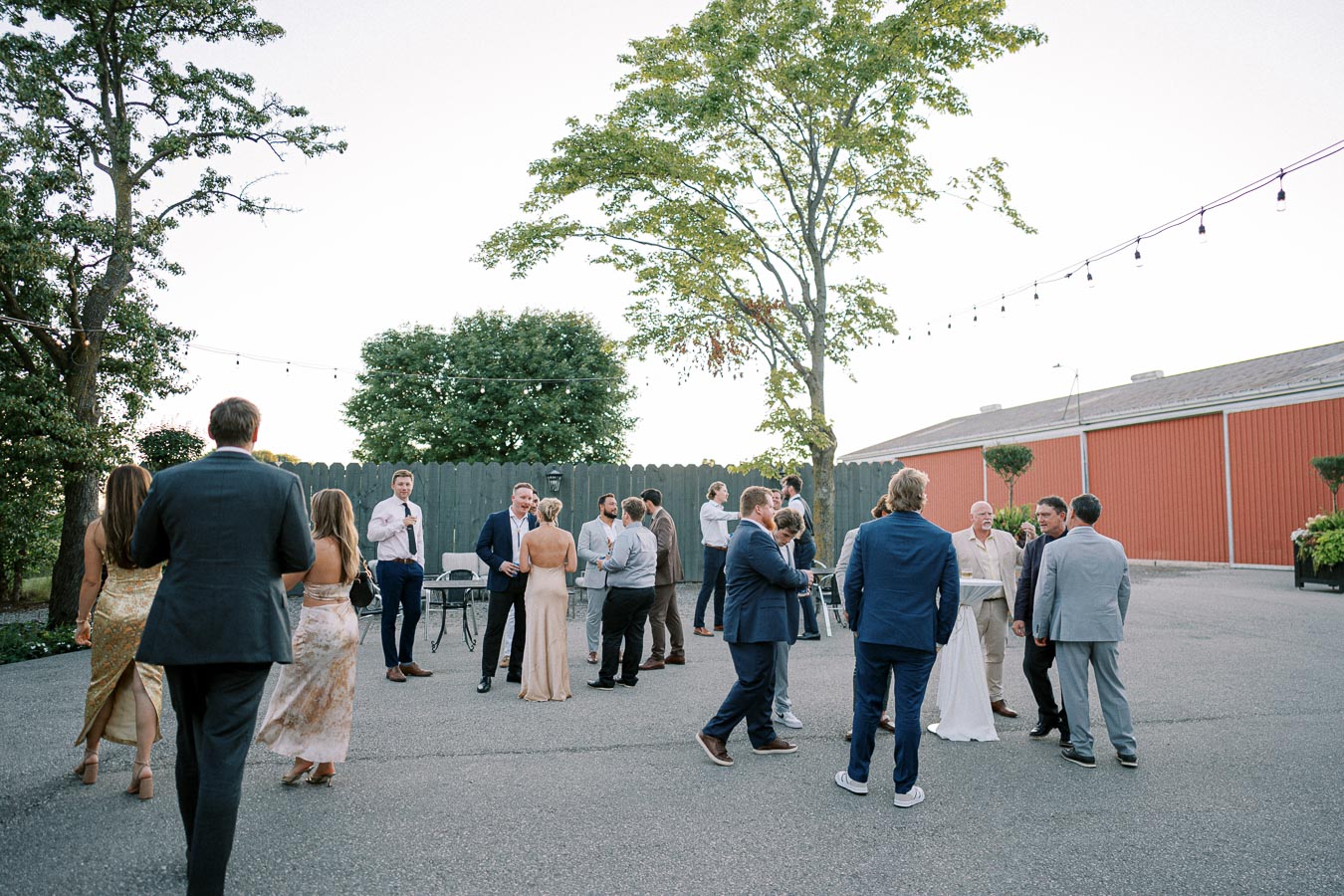 Outdoor wedding reception scene with guests mingling in formal attire, surrounded by greenery and decorative string lights, in front of a red barn on a clear day.