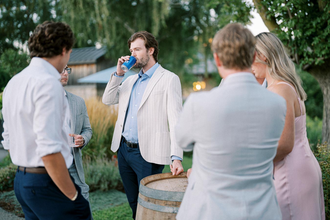 A group of well-dressed people socializing outdoors, with a man in a light striped jacket and blue shirt drinking from a can near a wooden barrel, surrounded by greenery and a rustic setting.