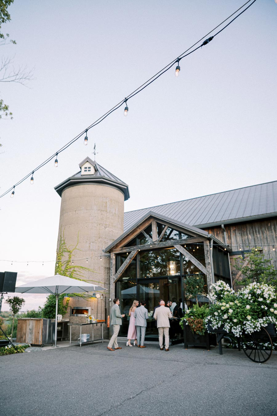 Guests gathering outside a rustic barn venue at sunset, featuring string lights and a historic silo with lush greenery, creating a charming countryside atmosphere.