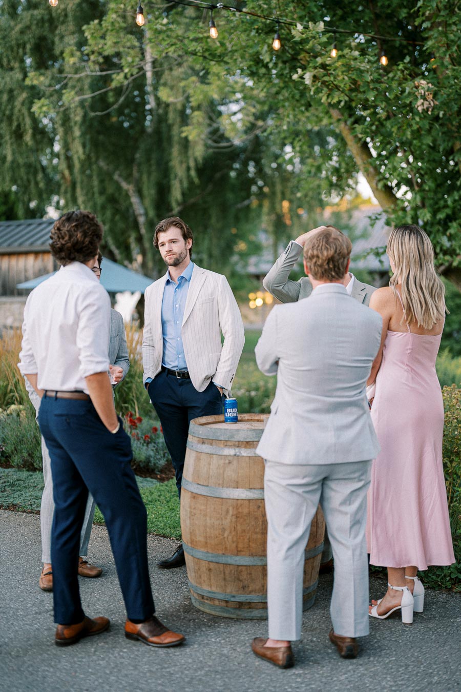 Group of elegantly dressed people gathered around a wooden barrel table at an outdoor event with string lights and lush greenery in the background.