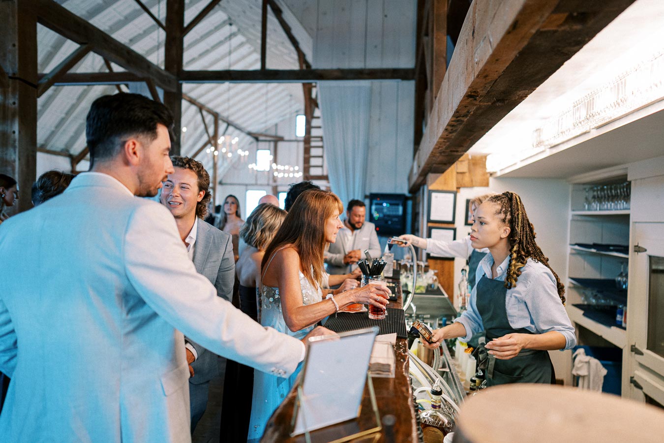 Bar setting with a group of well-dressed people socializing, receiving drinks from a bartender with braided hair, in a rustic venue with wooden beams and ambient lighting.