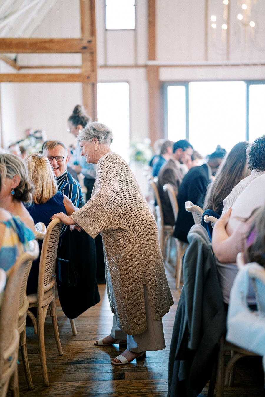 A lively gathering in a well-lit dining hall featuring people seated at tables in conversation, with a woman in a beige knitted cardigan interacting with guests, highlighting social interaction and communal joy.