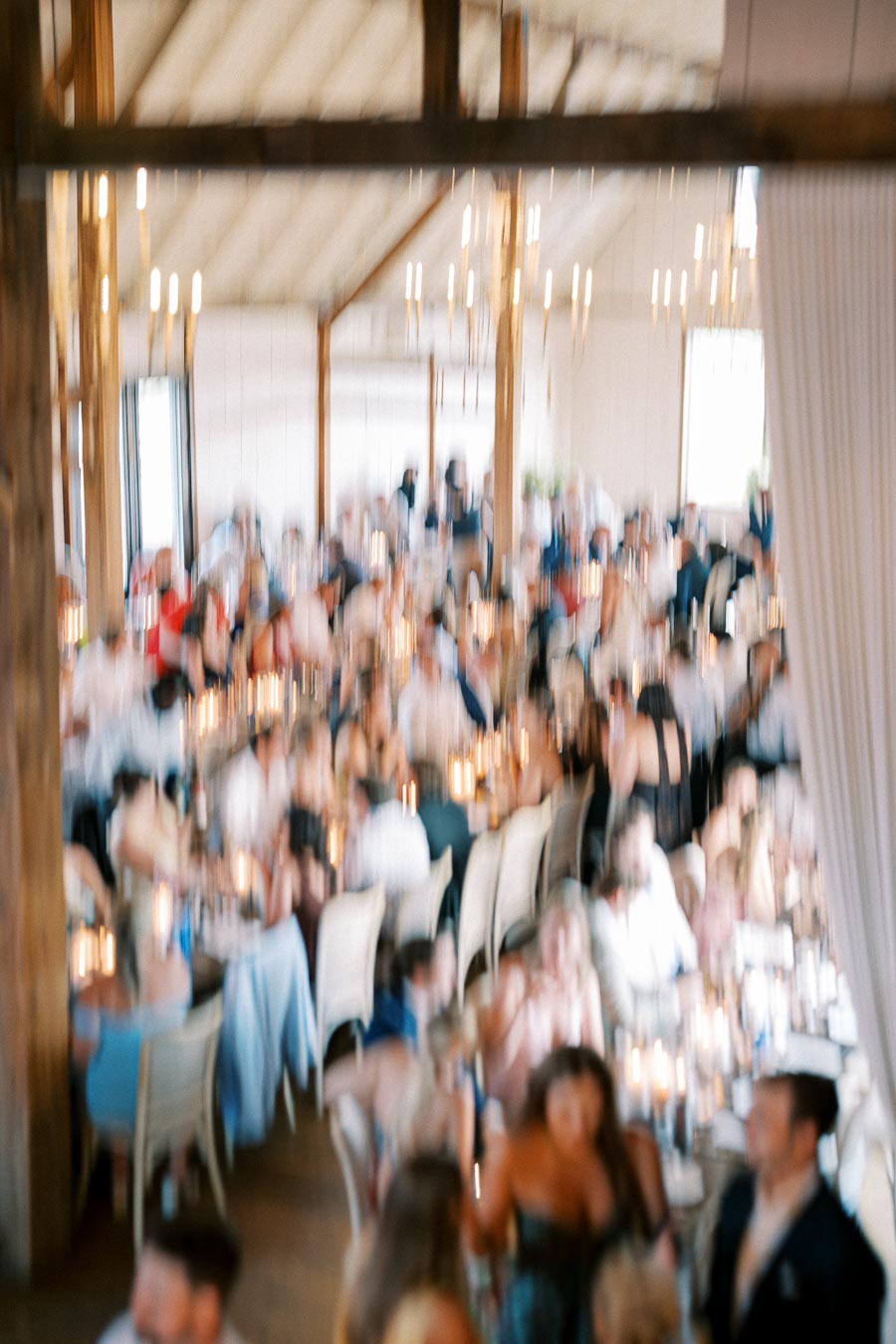 Blurry image of a crowded indoor event with people seated at tables under a high ceiling with chandeliers.