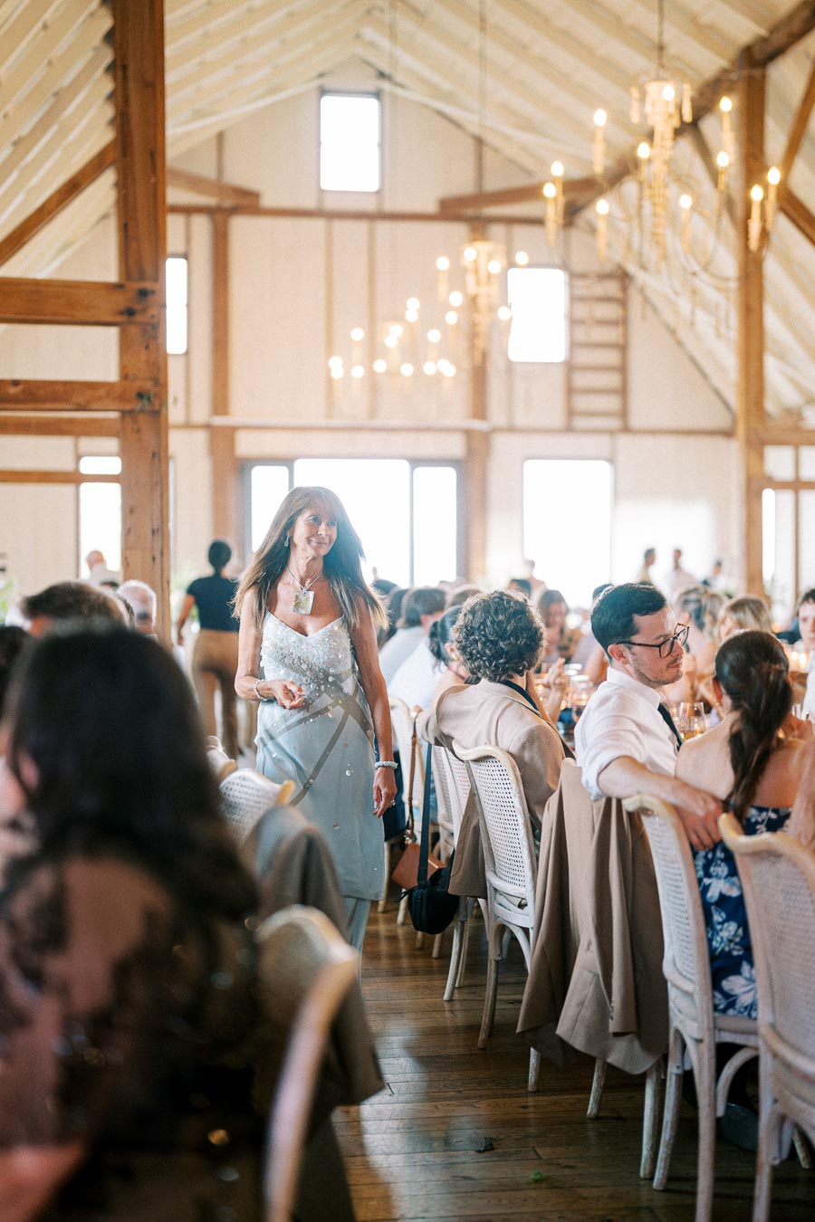 Woman in a light blue dress walking through a rustic, elegantly decorated wedding reception in a barn with chandeliers and guests seated on wooden chairs.