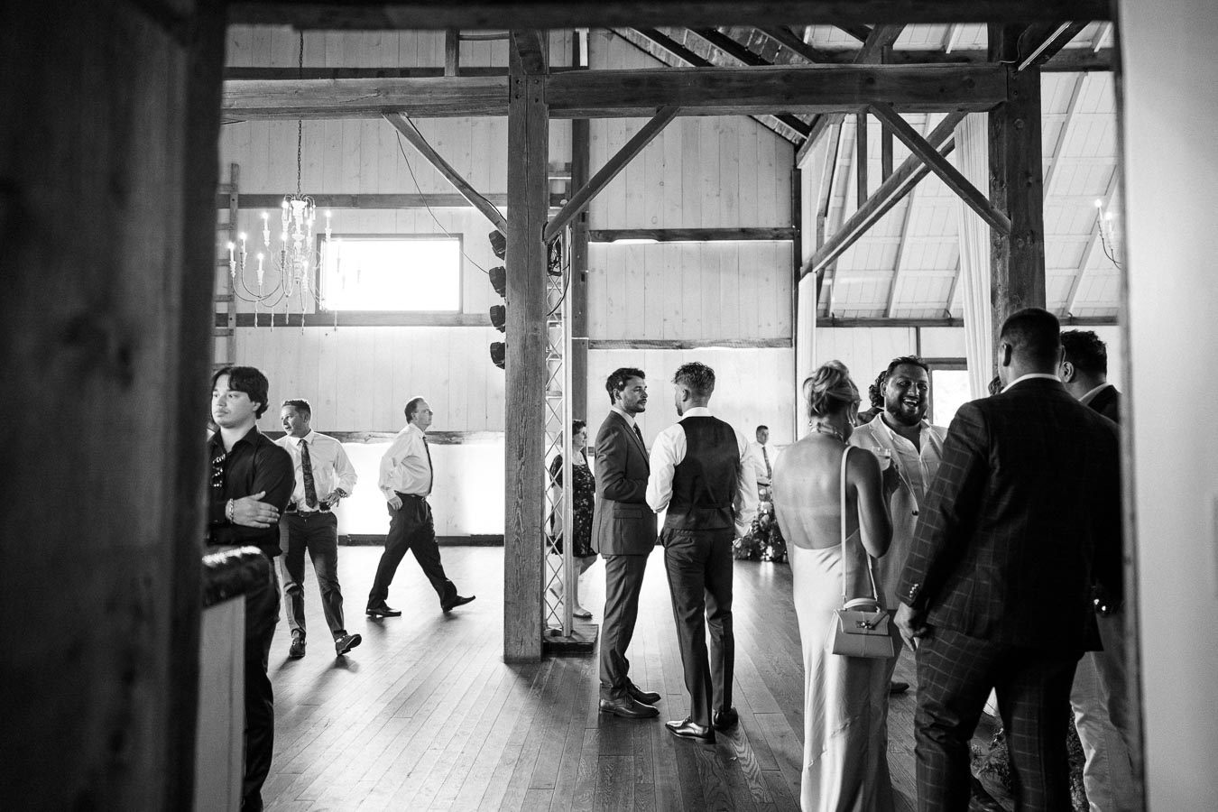 Black and white image of people socializing at a wedding reception in a rustic, barn-like venue. Natural light fills the wooden interior, highlighting guests dressed in formal attire, including suits and a gown, as they engage in conversations under a chandelier.