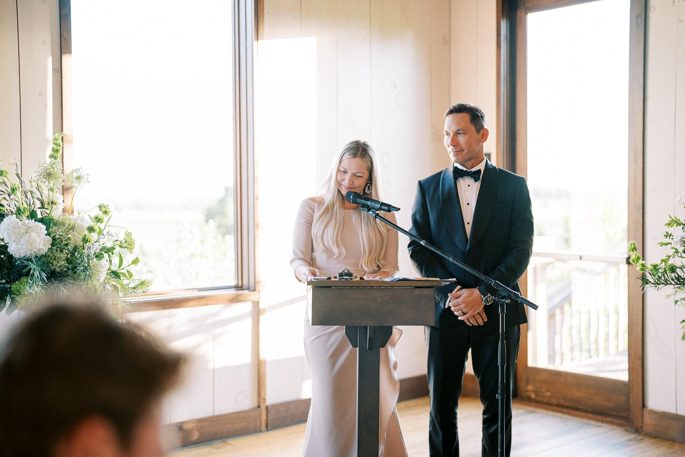 Elegant couple giving a speech at a wedding reception, with woman speaking at microphone and man in tuxedo standing beside her, indoors with large windows and floral decorations.