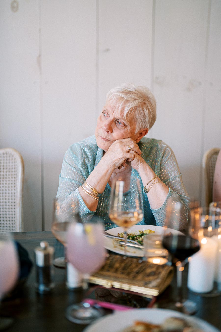 Elderly woman in a sparkly blue top sitting at a dining table with glassware and candles, looking thoughtfully to the side.