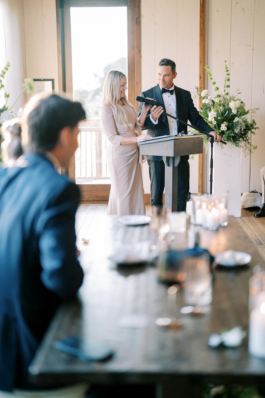 Elegant wedding reception scene with a man in a tuxedo and a woman in a formal dress giving a speech at a podium. Candles and floral arrangements decorate the bright, airy room, while seated guests listen attentively.