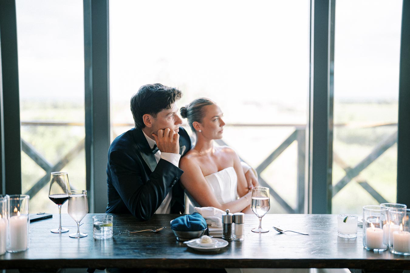 A bride and groom sitting thoughtfully at a reception table with wine glasses, candles, and a scenic window view in the background.