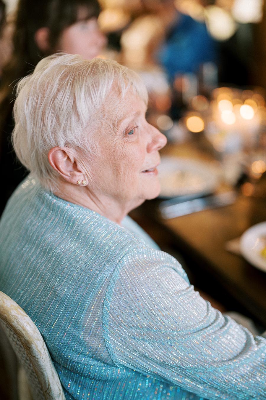 Elderly woman in a shimmering blue outfit sitting at a dining table, warmly lit background.