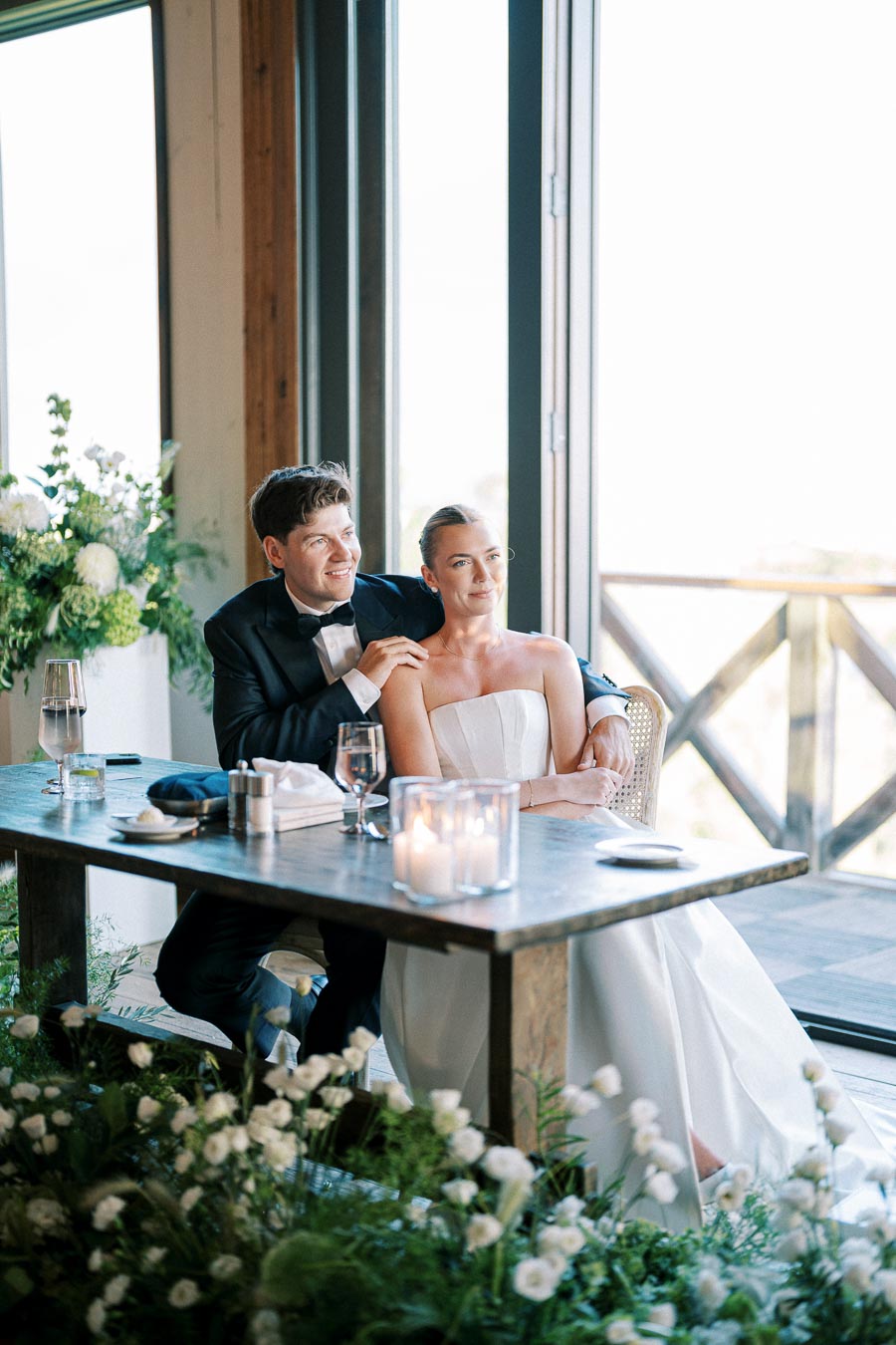 A bride and groom seated at a table by the window, elegantly dressed in wedding attire. The groom is in a black tuxedo and the bride in a strapless white gown, surrounded by green and white floral decorations. The table is set with candles, wine glasses, and dinnerware. Natural light filters through the large window, creating a serene and