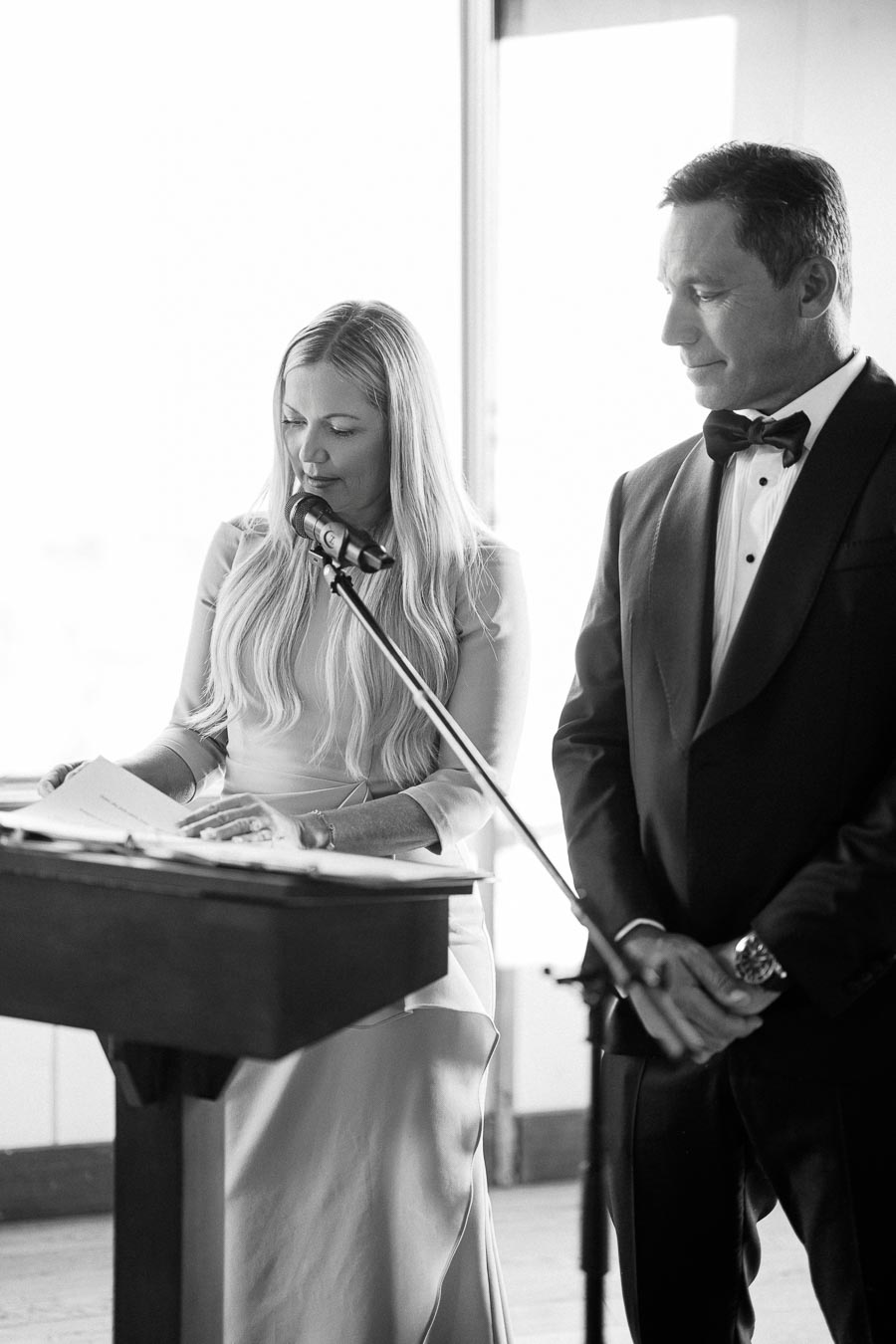 Elegant black and white photo of a woman reading a speech into a microphone at a podium, accompanied by a man in formal wear, possibly at a wedding or formal event.