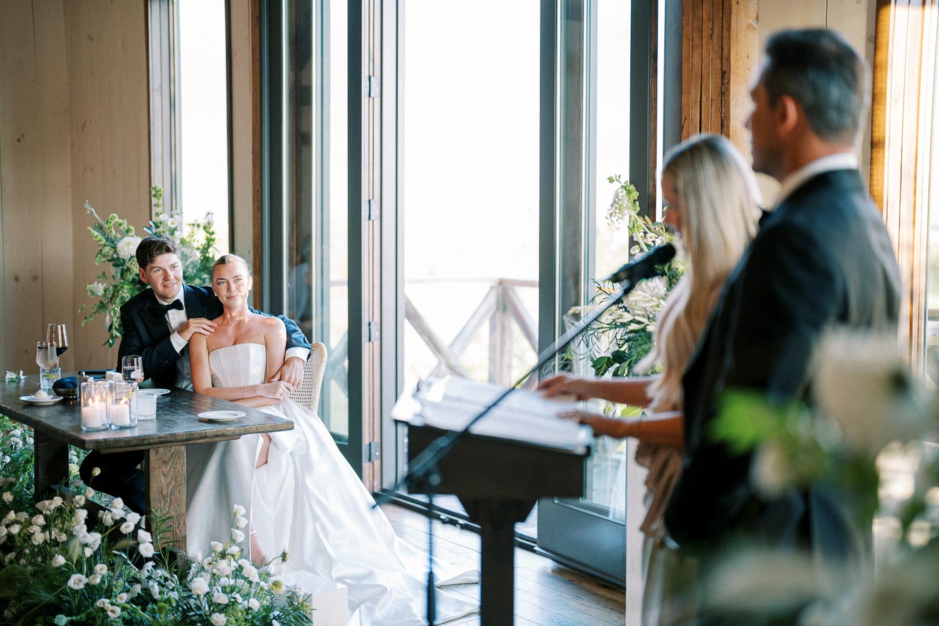 Elegant wedding scene with a bride in a strapless gown and groom in a tuxedo seated at a wooden table, listening to speeches. The room is decorated with lush flowers, candles, and large windows providing natural light. Guests in formal attire speak at a podium.