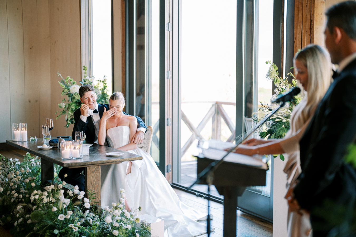 A bride and groom sitting at a decorated table, laughing during a wedding speech, with a speaker at a microphone and floral arrangements in a bright, airy venue.