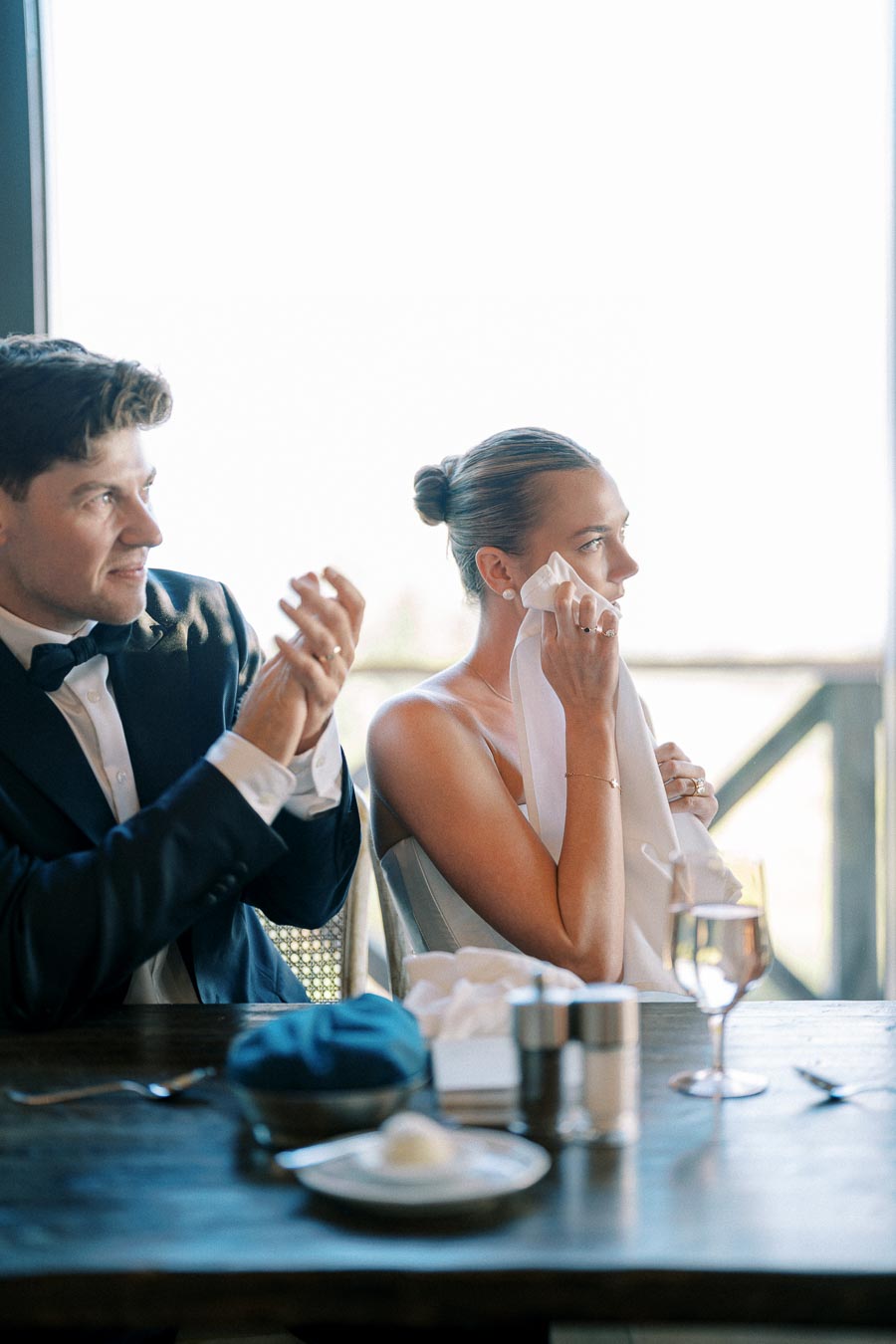Woman wiping tears with napkin next to man clapping, seated at elegant dinner event.