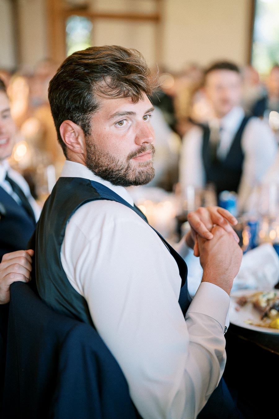 A man in a suit and tie sits at a table during a formal event, looking back over his shoulder.