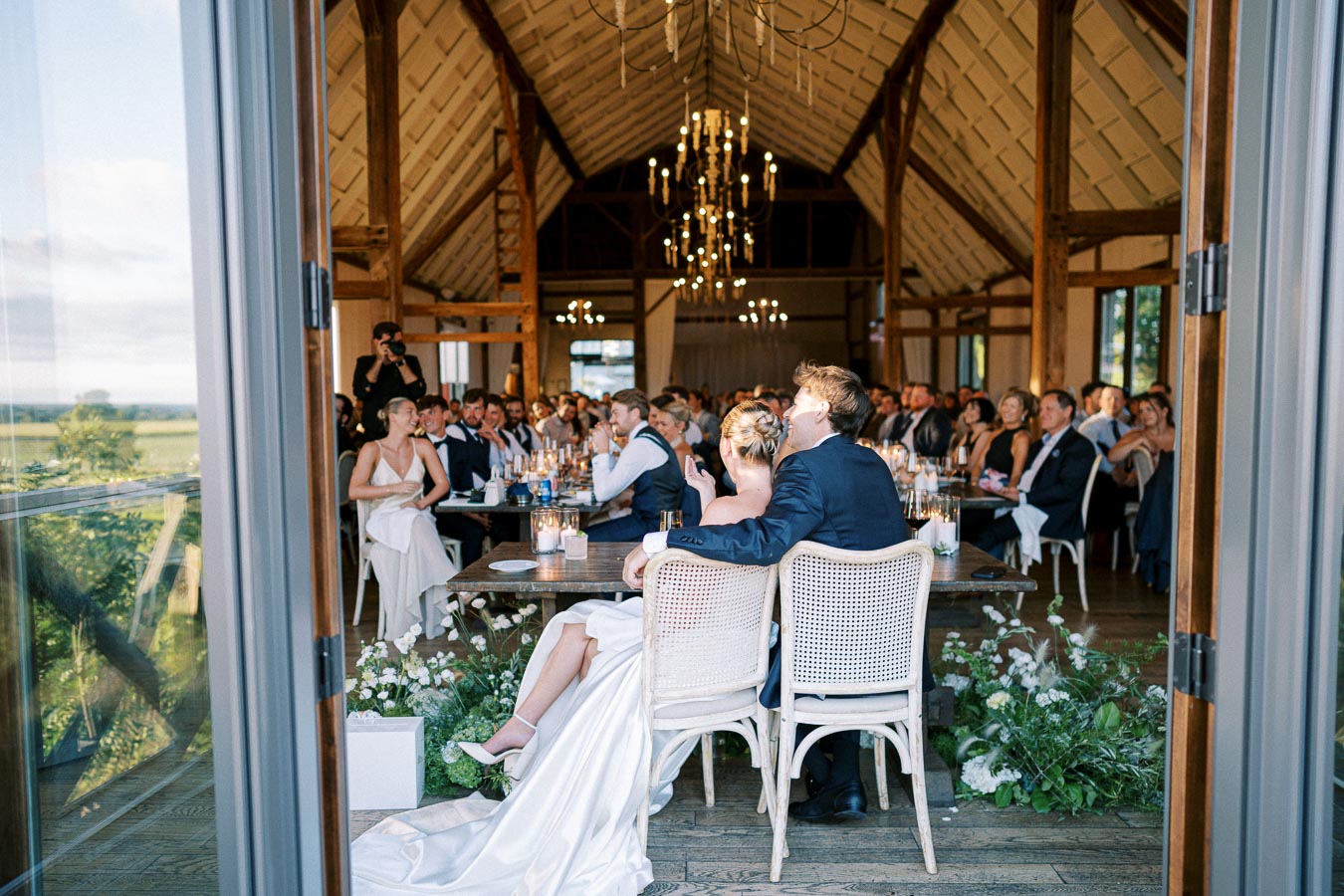 A bride and groom sit together at the head of a long dining table in a beautifully decorated barn during a wedding reception, surrounded by guests wearing formal attire. Chandeliers hang from the vaulted wooden ceiling, and flowers adorn the space.