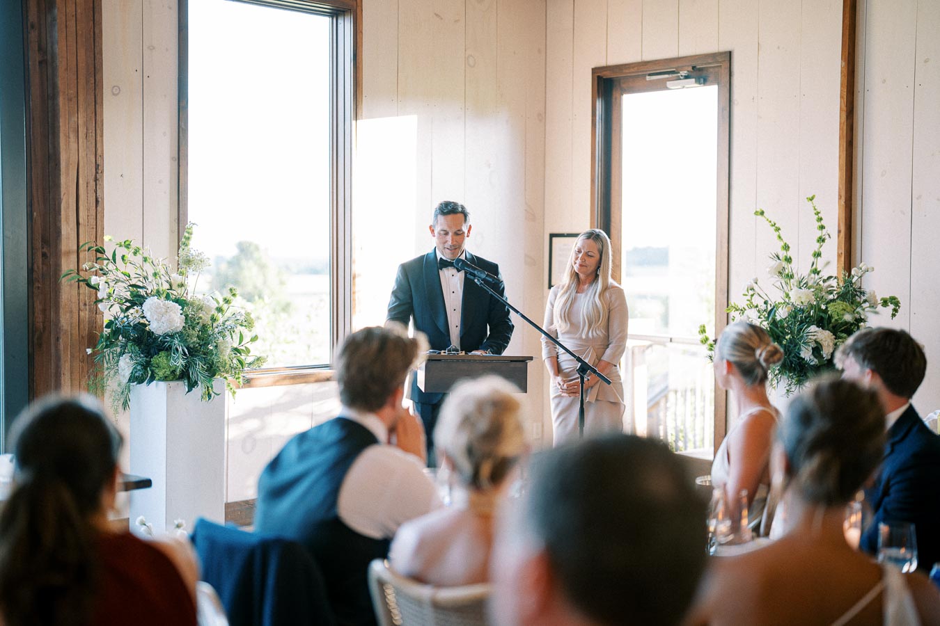 A man in a tuxedo delivers a speech at a podium with a microphone, while a woman in a light-colored dress stands by his side. They are in a well-lit room with large windows and decorative floral arrangements. Seated guests are visible in the foreground, suggesting a formal event or wedding reception.