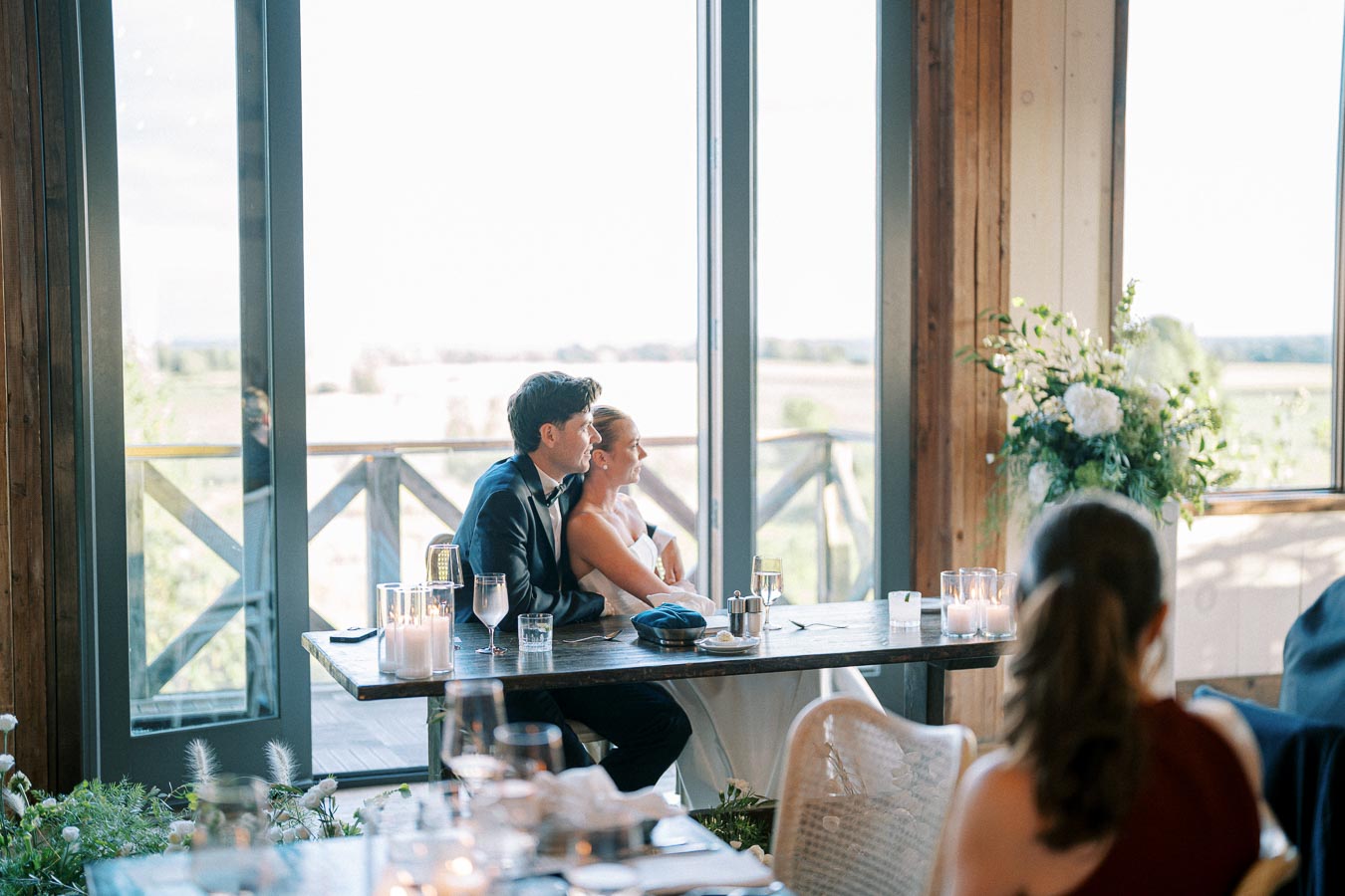 Wedding couple sitting at a reception table, embracing and gazing out large windows with scenic countryside views, surrounded by floral arrangements and candlelit glasses.