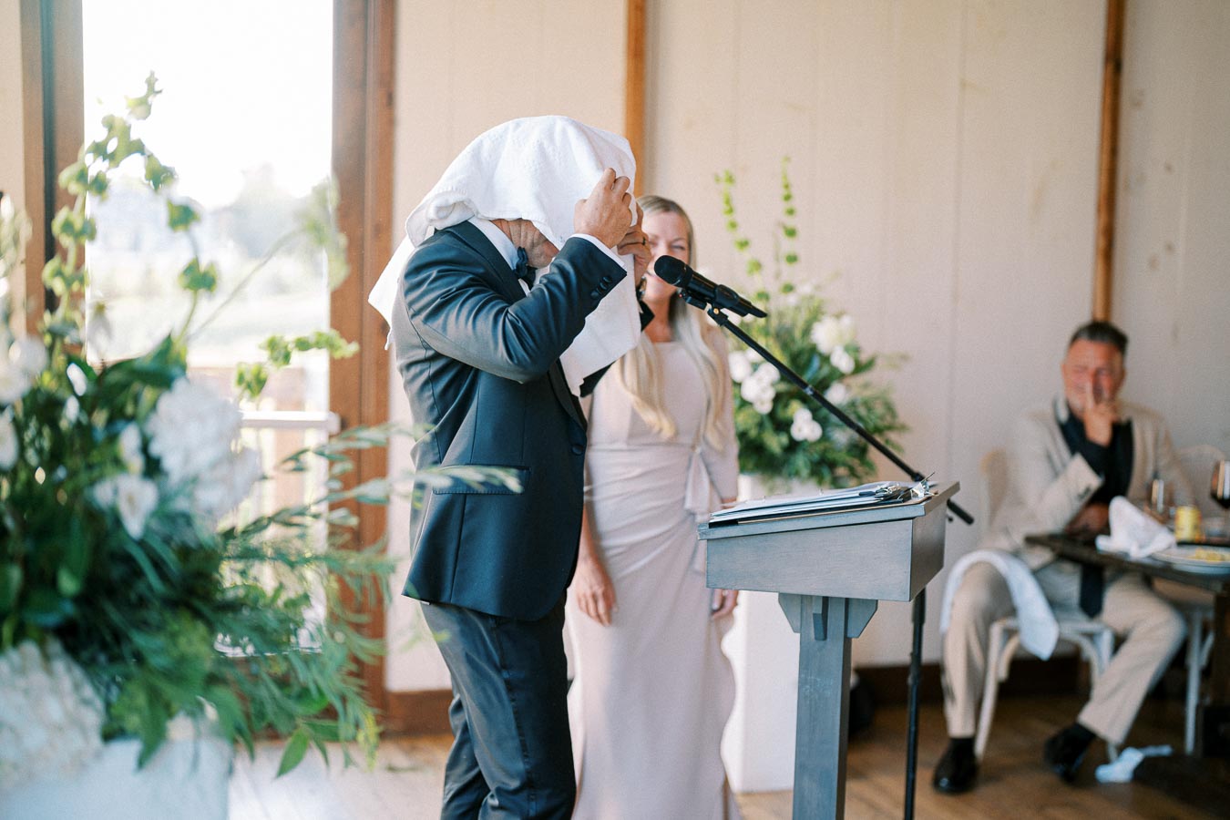 A man playfully covers his head with a napkin while standing at a podium during a wedding speech, with a woman smiling beside him in a floral-decorated venue.