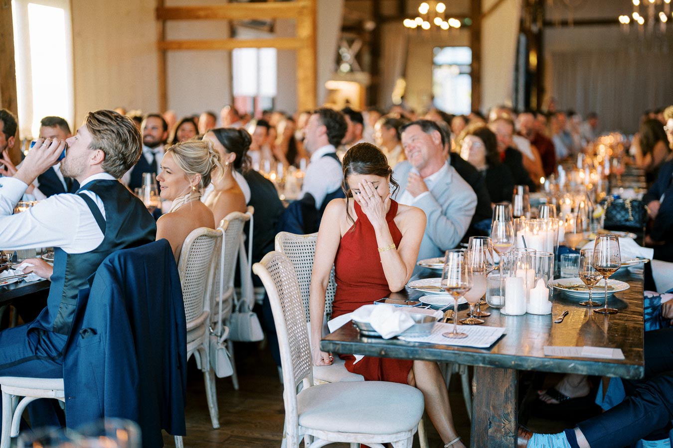 A lively wedding reception with guests seated at elegantly decorated tables, focused on a woman in a red dress smiling with her hand over her face, surrounded by laughing attendees and candlelit ambience.