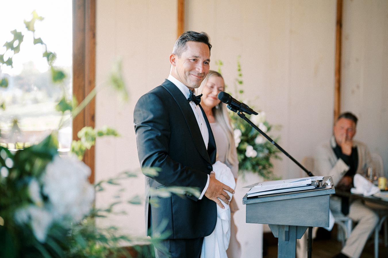 Wedding speaker in a tuxedo giving a heartfelt speech at a ceremony, with guests and floral decorations in the background.