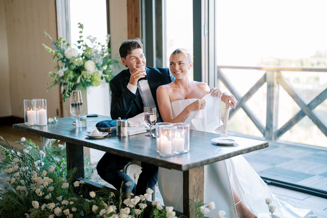 Wedding couple sitting at a table with elegant floral decorations and candle centerpieces, enjoying a moment together at a reception with a scenic view outside.