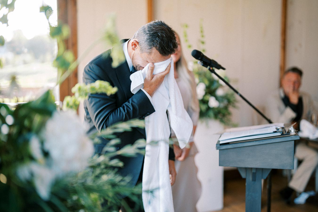 Man in a suit wiping tears with a white cloth during an emotional speech at a formal event, standing next to a microphone and surrounded by greenery and floral decorations.