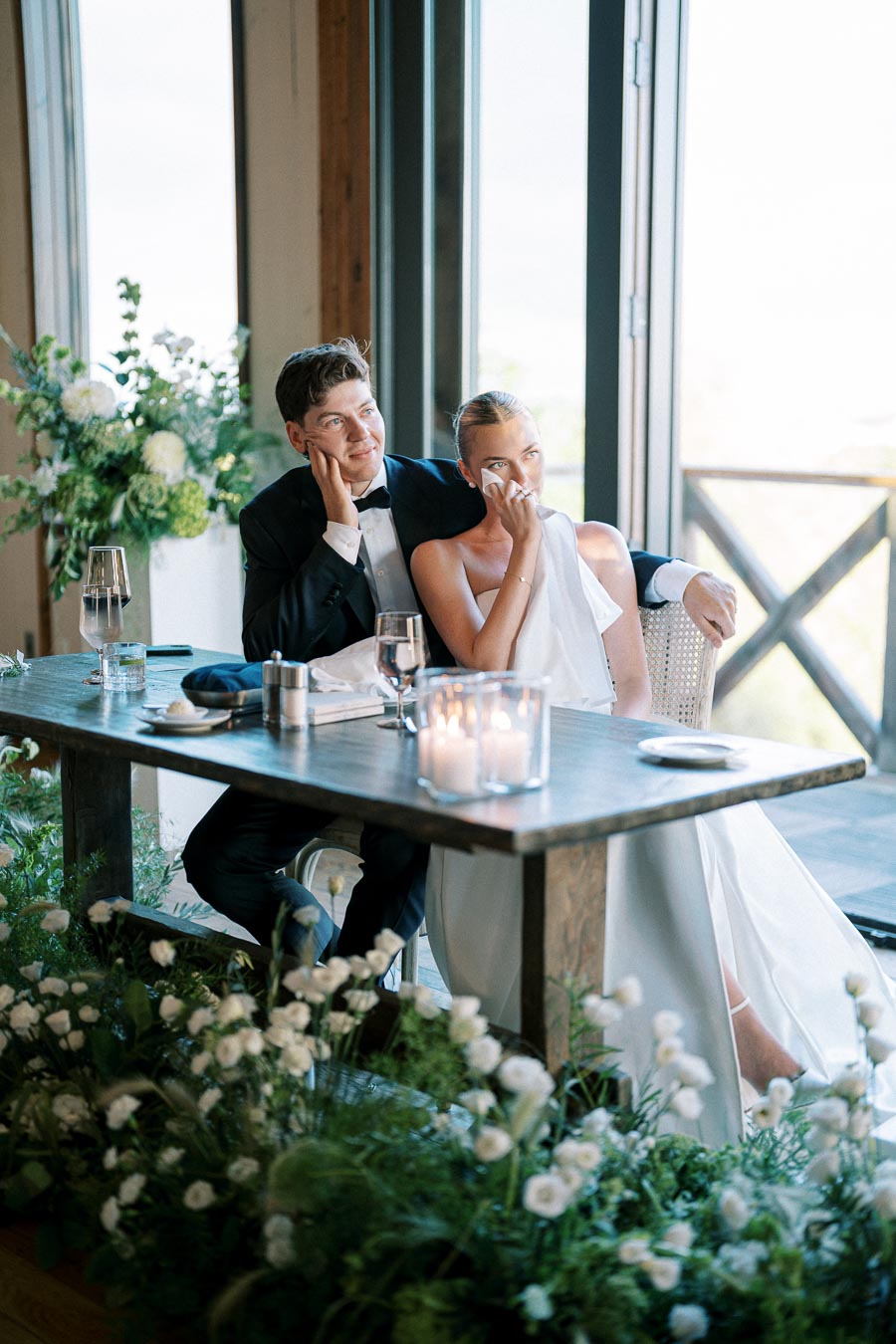 A bride and groom sit together at a beautifully decorated wedding reception table, surrounded by lush white flowers, with candles and glasses in the foreground. The couple appears emotional, gazing contentedly, capturing a heartfelt moment during their special day in a sunlit venue.