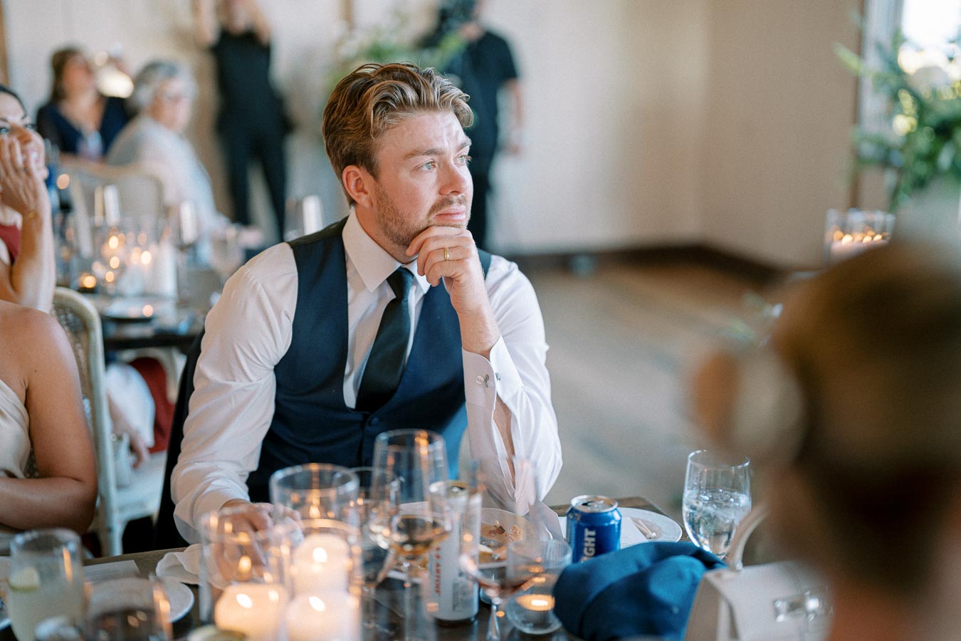 A man in a formal vest and tie sits thoughtfully at a candlelit event table, surrounded by guests, with a contemplative expression, in a softly lit room.