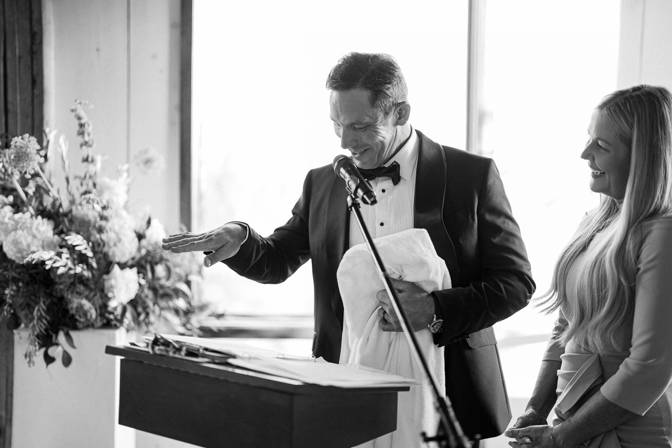 Black and white photo of a man in a tuxedo giving a speech at a podium, holding a napkin, with a smiling woman standing beside him.