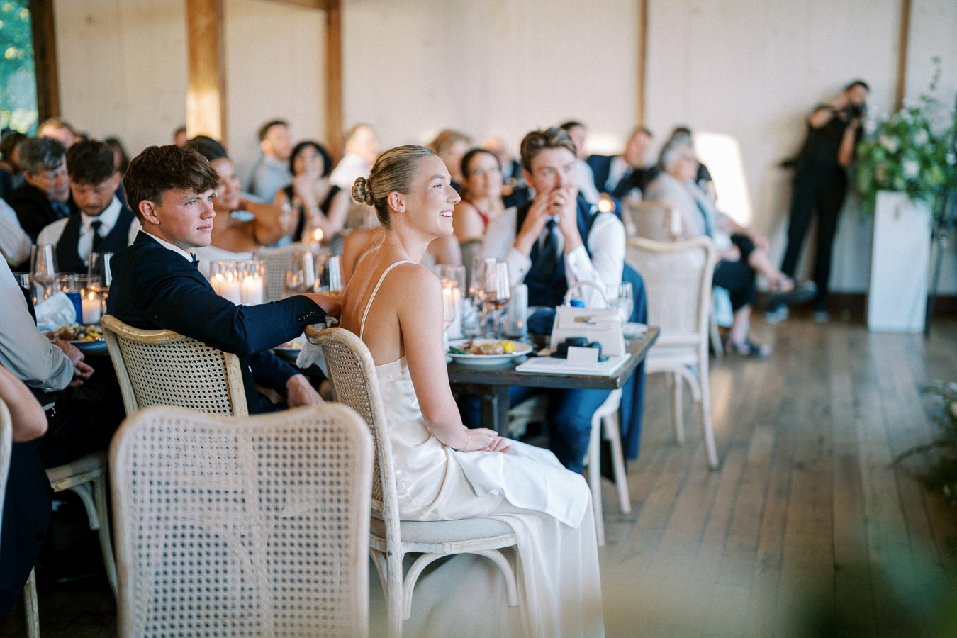 Woman in a white dress smiling at a dining table during a wedding reception, surrounded by guests in formal attire, with candles and tableware creating an elegant atmosphere.