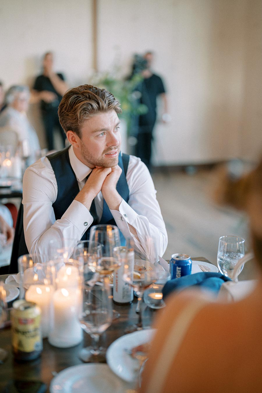 A man in formal attire sitting at a candlelit table, looking thoughtfully into the distance during a wedding reception.